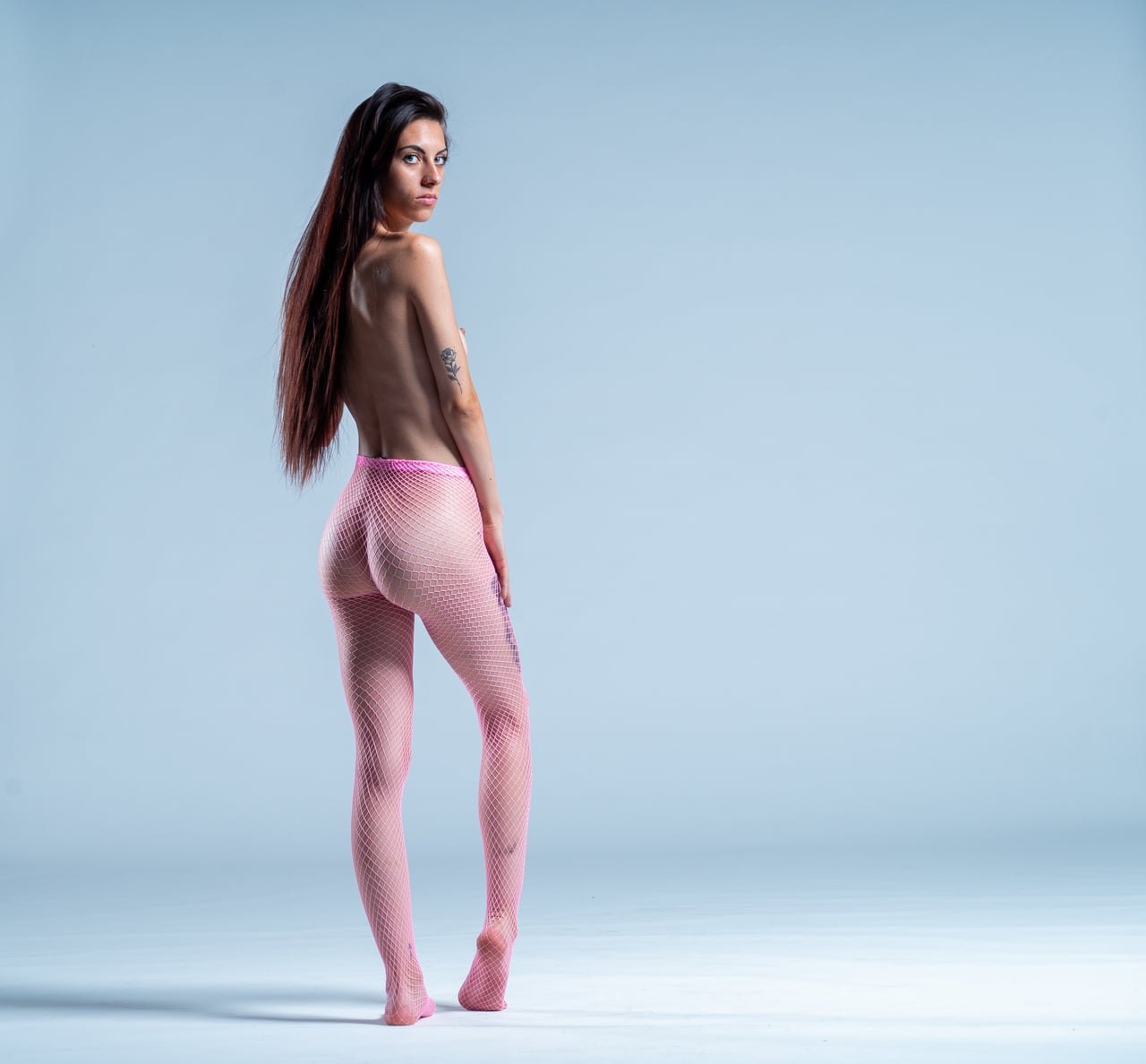 Dona d'esquena amb mitges de malla rosa transparent cobrint-li de cintura a peus, tors nu, mirada per damunt de l'espatlla, fons d'estudi blau grisenc.