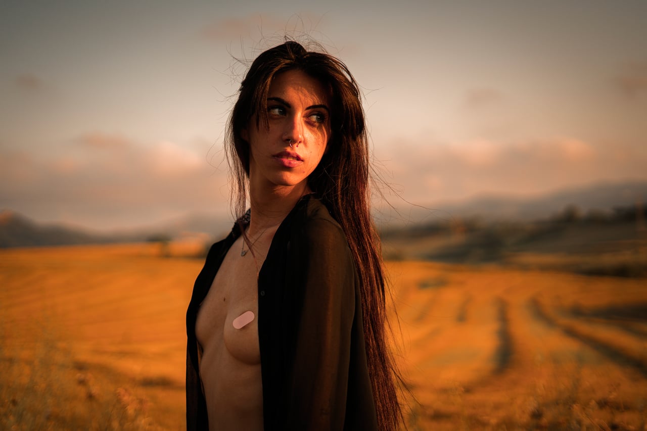 Retrat editorial de dona amb camisa oberta deixant un pit parcialment cobert, paisatge rural de turons al capvespre al darrere.