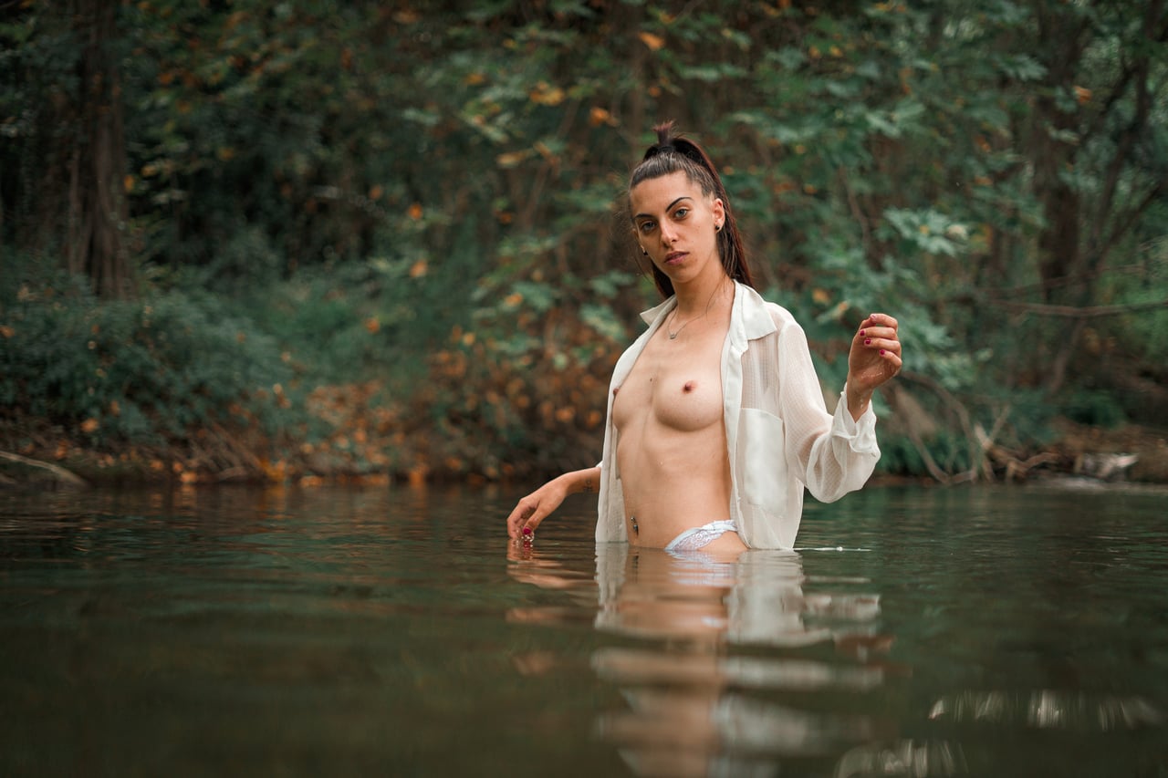 Dona dempeus amb l'aigua fins a la cintura, camisa blanca oberta sobre el tors nu, mirada directa a càmera, rierol del bosc al fons.